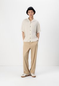 Light beige short-sleeve shirt with brown buttons, paired with beige wide-leg trousers and white sneakers with brown accents. Black bucket hat.