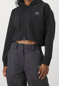 Černá zkrácená mikina s kapucí na zip a nastavitelnou kapucí, s malým kulatým logem. V kombinaci se šedými kalhotami, které jsou u pasu těsné.