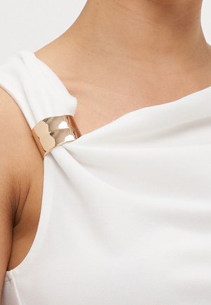 Gros plan d'un haut blanc sans manches avec un détail d'anneau doré brillant tenant le tissu à l'épaule près de la clavicule.