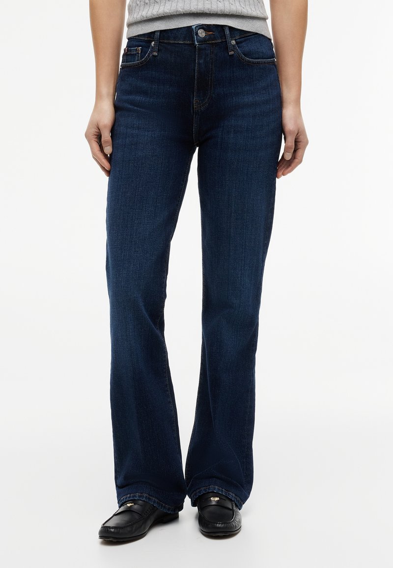 Personne portant un jean bootcut bleu foncé, un pull côtelé gris clair et des mocassins noirs, debout devant un fond blanc.