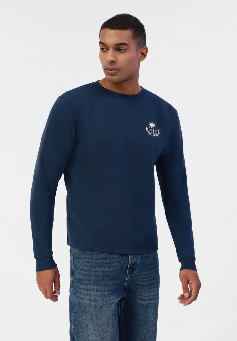Sweatshirt bleu marine à manches longues en mélange de coton avec col rond, présentant un petit logo blanc de palmier sur la poitrine et des poignets côtelés.