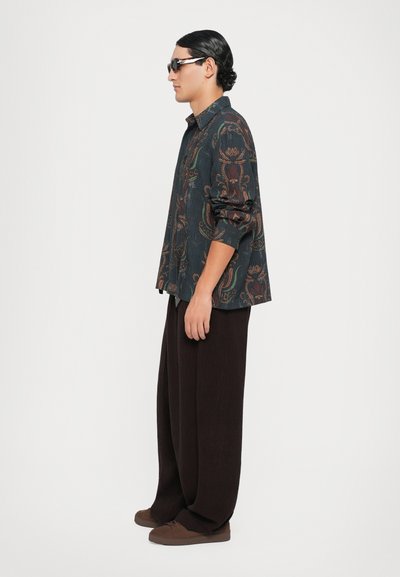 Camicia a maniche lunghe con complessi motivi floreali multicolori su uno sfondo scuro, abbinata a pantaloni marroni larghi e scarpe scure.