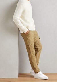 Pull en laine ivre, pantalon cargo kaki avec poches latérales et baskets blanches. Les textures douces et les couleurs muted créent un look décontracté.