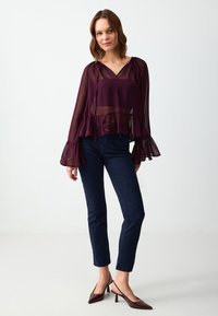 Blusa trasparente bordeaux con scollatura a V, orlo arricciato e maniche a campana, abbinata a pantaloni attillati blu scuro e scarpe col tacco color mogano.