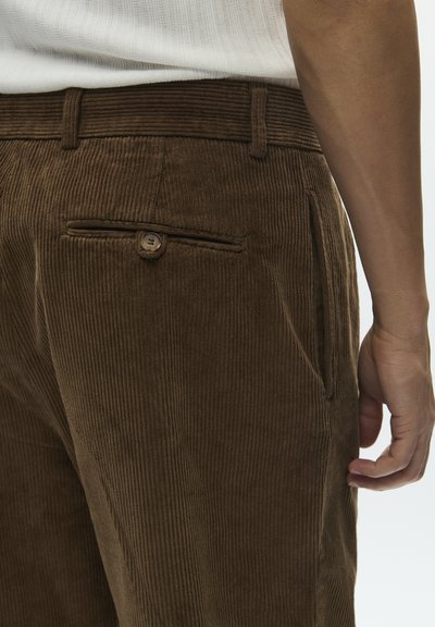 Close-up of a person wearing brown corduroy pants with a buttoned back pocket and a white textured shirt tucked in.