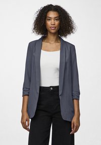 Námornícky blazer s hladkým materiálom, otvoreným predným dielom a mierne odhodenými rukávmi. Nosený cez biely top, v kombinácii s čiernymi nohavicami s vysokým pásom.
