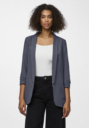 Navy blazer with a smooth fabric, open front, and slightly rolled sleeves. Worn over a white top, paired with high-waisted black pants.