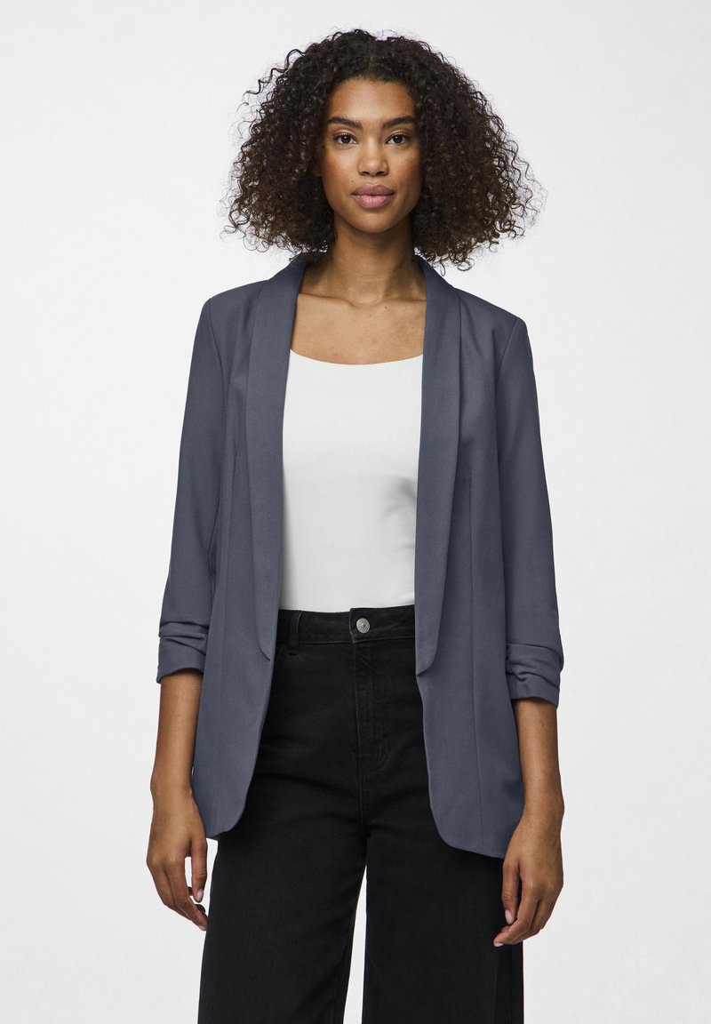 Námornícky blazer s hladkým materiálom, otvoreným predným dielom a mierne odhodenými rukávmi. Nosený cez biely top, v kombinácii s čiernymi nohavicami s vysokým pásom.