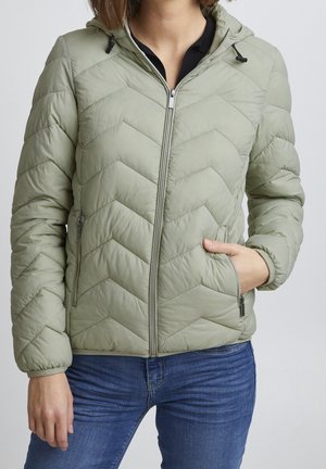 Femme portant une veste matelassée vert clair zippée avec capuche et un jean bleu, une main dans la poche de la veste sur fond uni.