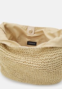 Sac rond tressé en matière beige semblable à de la paille, avec un intérieur en tissu beige lisse. Il possède une fermeture pression et une étiquette de marque noire à l'intérieur.