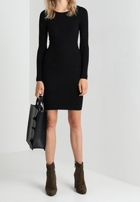Robe longue noire en maille avec manches longues et silhouette ajustée. Associée à des bottines vert olive et un sac  à main structuré noir.