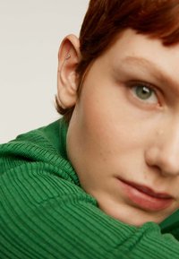 Primer plano de la cara de una persona con cabello corto castaño rojizo, que lleva un suéter verde texturizado y tiene un pequeño pendiente de perno.