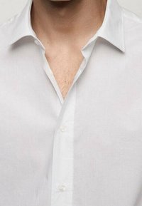 Biała koszula zapinana na guziki o gładkiej strukturze, klasycznym kołnierzyku i małych przezroczystych guzikach, które odsłaniają fragment skóry przy dekolcie.