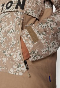 Kurtka w wzór camouflage z zapięciem na zamek, w odcieniach beżu i zieleni, z rzepami na rękawach oraz ściągaczem u dołu.