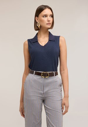 Donna con capelli castani lisci indossa un top senza maniche blu navy, pantaloni a righe a vita alta, cintura marrone e un grande orecchino dorato a forma di foglia, guarda verso destra.