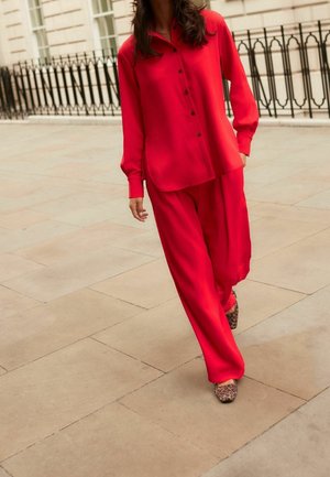 Kvinde går på flisebelagt fortov iført en løs rød skjorte med knapper, matchende vidde bukser og sko med leopardprint.