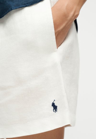 Χέρι στην τσέπη, φοράει λευκά βερμούδα από λινό με λογότυπο παίκτη πόλο σε ναυτικό μπλε κοντά στο τελείωμα.