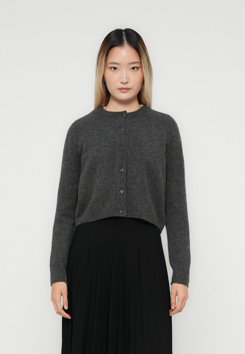 Woman with straight ombre hair wearing a dark gray buttoned cardigan and a black pleated skirt, standing against a plain light background.