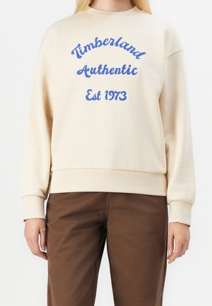 Person iført en cremefarvet sweatshirt med blå tekst "Timberland Authentic Est 1973" og brune bukser, stående mod en hvid baggrund.