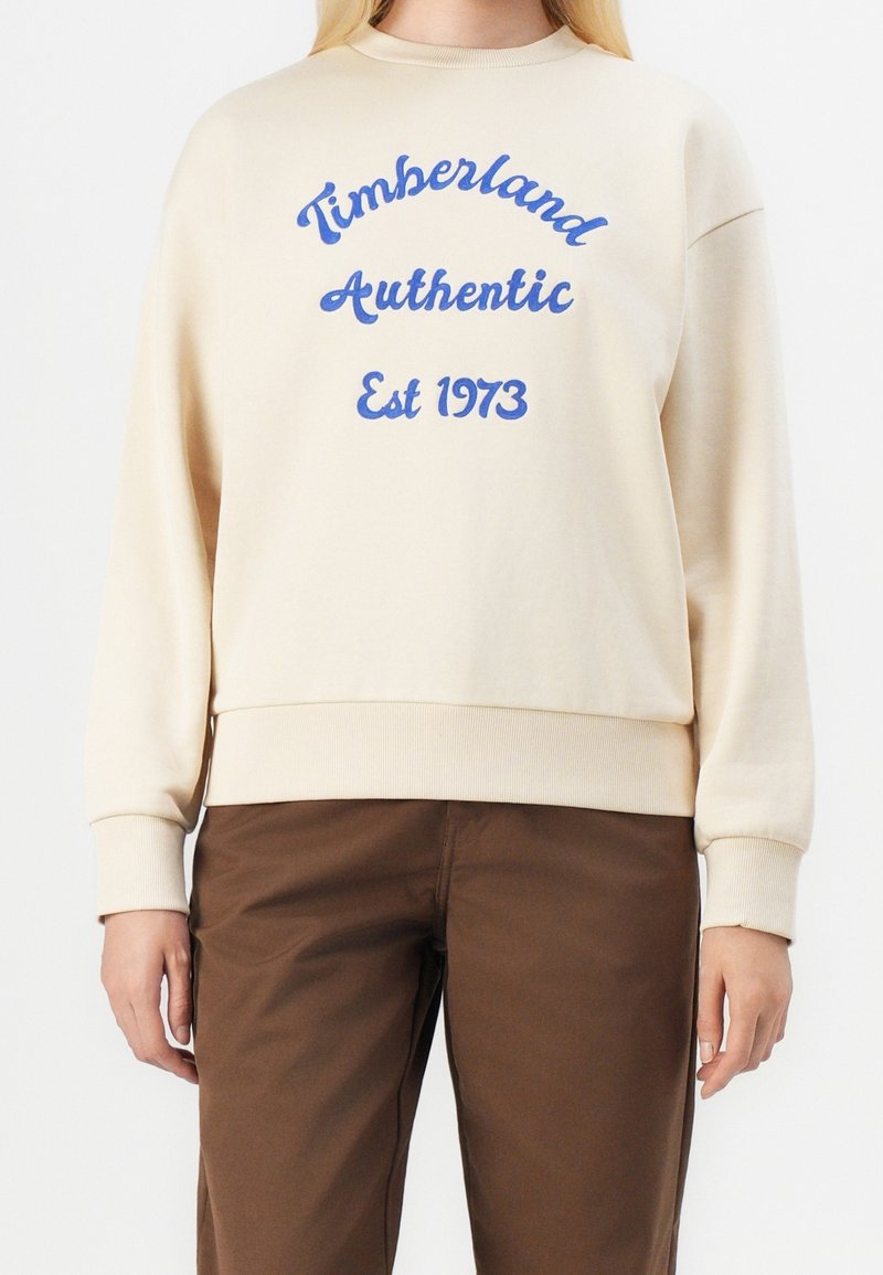 Personne portant un sweat crème avec le texte bleu « Timberland Authentic Est 1973 » et un pantalon marron, debout devant un fond blanc.