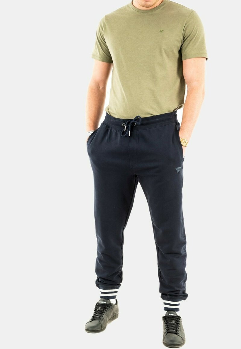 Camiseta de manga corta verde oliva y pantalones de chándal azul marino con cintura elástica y puños de rayas blancas, combinados con zapatillas negras.