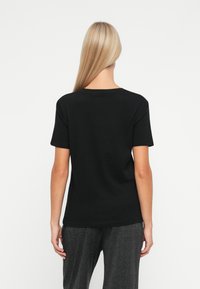 Kvinde med langt blondt hår iført en ensfarvet sort kortærmet t-shirt og grå bukser, stående med ryggen til mod en hvid baggrund.