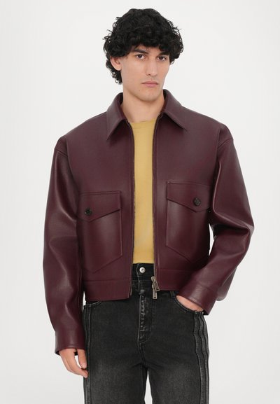 Burgunderfarbene, aus Kunstleder gefertigte kurze Jacke mit einem breiten Kragen, zwei vorderen Taschen und einem Reißverschluss. Gelegt über einem senfgelben Hemd.
