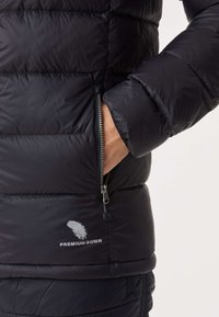 Svart puffjacka med quiltade sektioner, ficka med dragkedja och "PREMIUM DOWN"-logotyp. Slät, glänsande tyg med en texturerad yta.