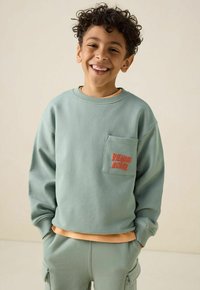 Sweatshirt vert menthe avec une poche avant et des broderies corail, assorti à un pantalon de survêtement, offrant une texture douce et une coupe décontractée.