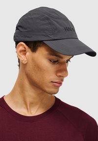 Dunkelgraue Mütze mit einem gebogenen Schirm, aus strukturiertem Stoff und mit gesticktem "HALO"-Logo. Getragen von einem Modell in einem kastanienbraunen Shirt.