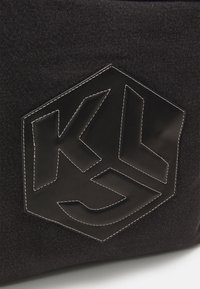Bolso de denim negro con un parche de cuero hexagonal que presenta un logo troquelado y costuras blancas en contraste. Diseño estructurado con materiales texturizados.