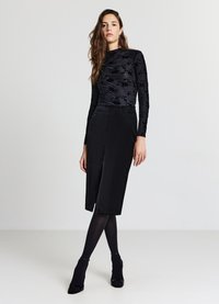 Schwarzes Langarmshirt mit strukturiertem Blumenmuster, kombiniert mit einem knielangen schwarzen Rock mit Frontschlitz, ergänzt durch schwarze High Heels.