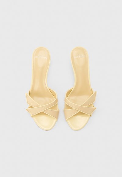 Μπεζ σανδάλια χωρίς λουράκι με σταυρωτές φαρδιές λουρίδες, ανοιχτά δάχτυλα και επίπεδες σόλες, σε θέα από πάνω σε απλό φόντο.