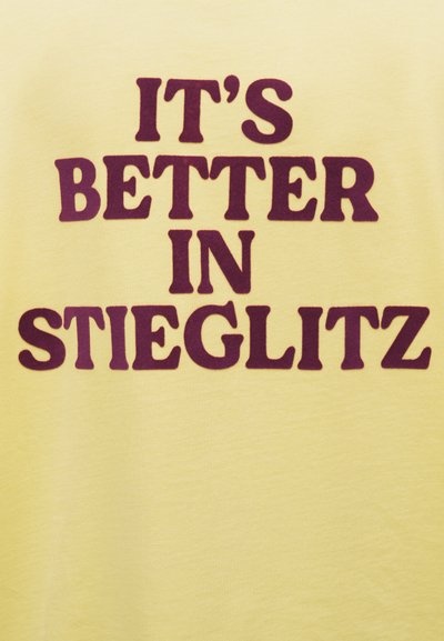Κίτρινο ύφασμα με έντονο σκούρο κόκκινο κείμενο που γράφει «ΕΙΝΑΙ ΚΑΛΥΤΕΡΑ ΣΤΟ STIEGLITZ», τοποθετημένο στο κέντρο.