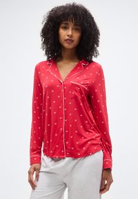 Κόκκινο σατέν τοπ πιτζάμας με λευκό τύπωμα φιόγκου, διαθέτει τσέπη, μακριά μανίκια και αντίθετη λευκή διακόσμηση στο γιακά και στο μέρος με τα κουμπιά.