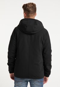 Veste matelassée noire avec capuche, dotée de poignets élastiques, d'une texture lisse et de détails de couture subtils. Portée avec un jean bleu.