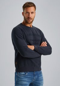 Tamno plava dukserica s reljefnim natpisom sprijeda, okruglim izrezom i rebrastim manžetama. Upari s svijetloplavim trapericama.