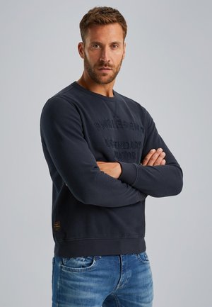 Donkerblauwe sweatshirt met verhoogde letters op de voorkant, ronde halslijn en geribde manchetten. Gecombineerd met lichtblauwe jeans.