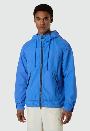 Hombre que lleva una chaqueta con capucha de cremallera en azul brillante con bolsillos delanteros y pantalones color crema, de pie frente a un fondo neutro.
