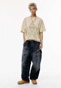 Beige, oversized t-shirt zöld grafikai designnal, lazán illeszkedő sötét farmer munkanadrággal és világosbarna bokacsizmával egy sima háttér előtt.