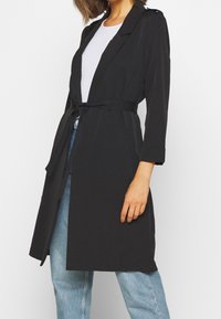 Trench-coat léger noir avec taille ceinturée, revers crantés, poches latérales et manches trois-quarts, associé à un jean bleu clair.