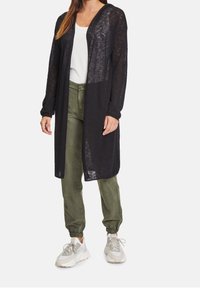 Černý dlouhý kardigan s lehkým, průsvitným materiálem, otevřeným předním dílem a dlouhými rukávy, kombinovaný s olivově zelenými kalhotami a bílými teniskami.