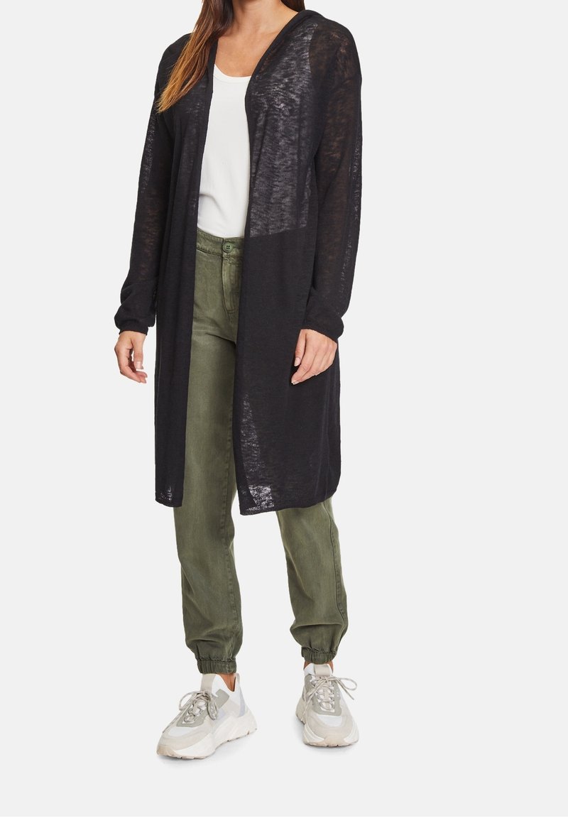 Černý dlouhý kardigan s lehkým, průsvitným materiálem, otevřeným předním dílem a dlouhými rukávy, kombinovaný s olivově zelenými kalhotami a bílými teniskami.