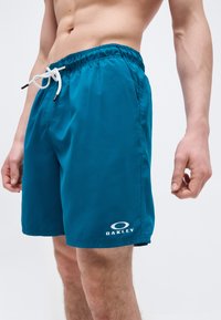 Αρσενικός κορμός και πόδια που φορούν μπλε μαγιό Oakley με λευκό κορδόνι, στέκονται μπροστά σε ένα απλό ανοιχτό φόντο.