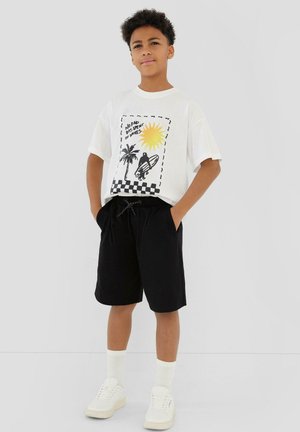 Garçon debout, les mains dans les poches, portant un t-shirt blanc avec un motif soleil, palmier, surfeur et un short noir sur fond gris.