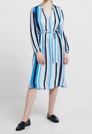 Femme portant une robe mi-longue à manches longues avec des rayures verticales en bleu, noir et blanc, associée à des mocassins noirs.