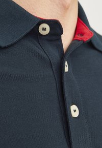 Tmavě námořnická polo trička s texturovaným úpletem, červeným detailem límce a zaoblenými plastovými knoflíky. Detailní záběr ukazuje detaily límce a knoflíků.