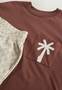 Donkerrood katoenen T-shirt met een beige, gestreepte onderkant; met een witte geborduurde palmboom op het borstzakje. Zachte textuur, casual ontwerp.