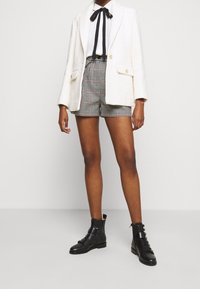 Witte gestructureerde blazer, zwarte vlinderdas, gemusterde shorts met rode accenten, en zwarte enkellaarzen met gespen. Eenvoudige, moderne uitstraling.
