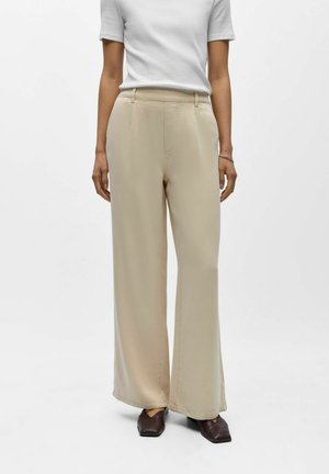 Kobieta w beżowych spodniach z szerokimi nogawkami, białej bluzce z krótkim rękawem i ciemnobrązowych butach z kwadratowym noskiem, stojąca na jednolitym tle.